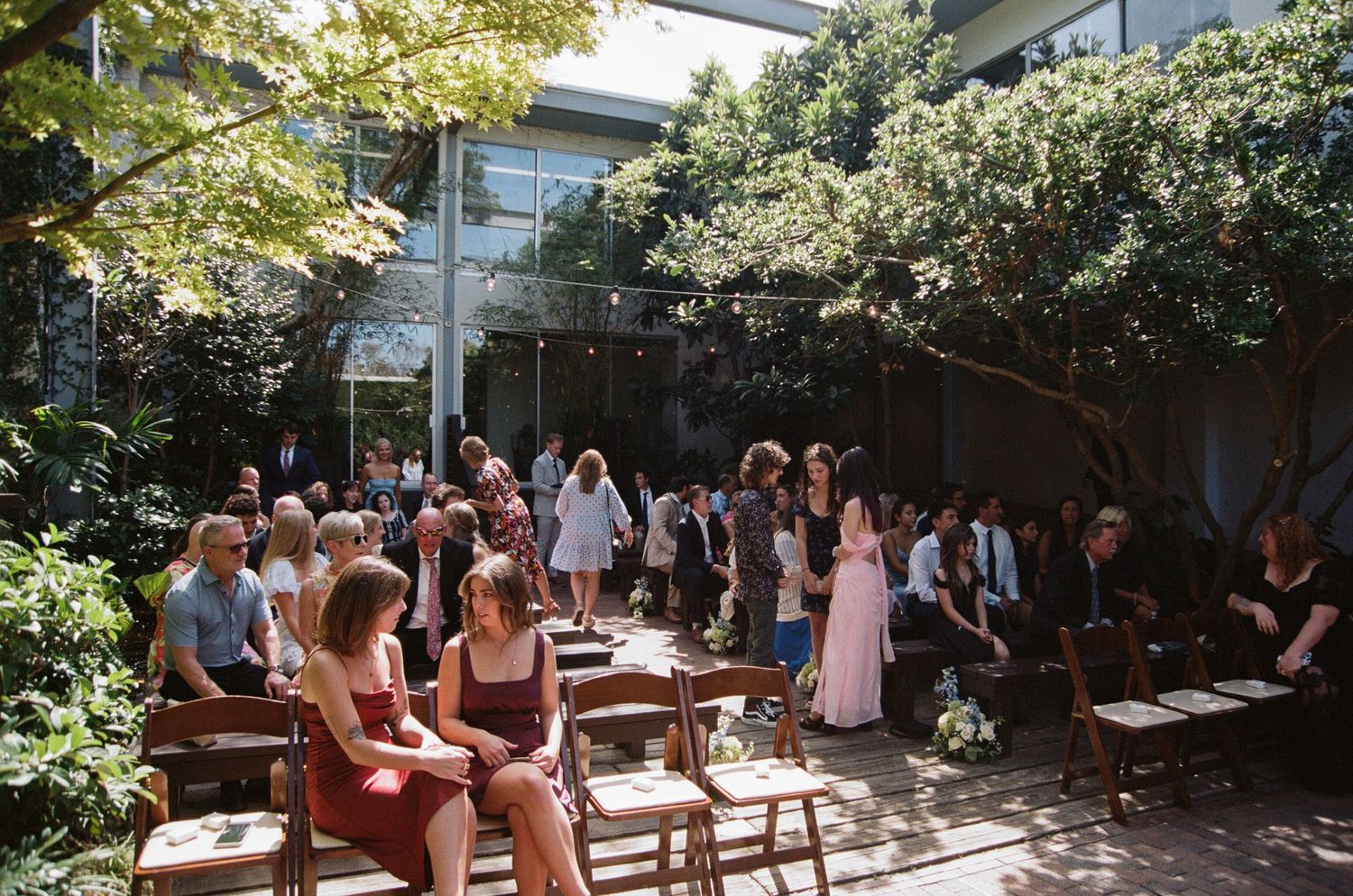 People enjoying outdoor dining in a sunny garden patio with wooden tables and lush greenery.