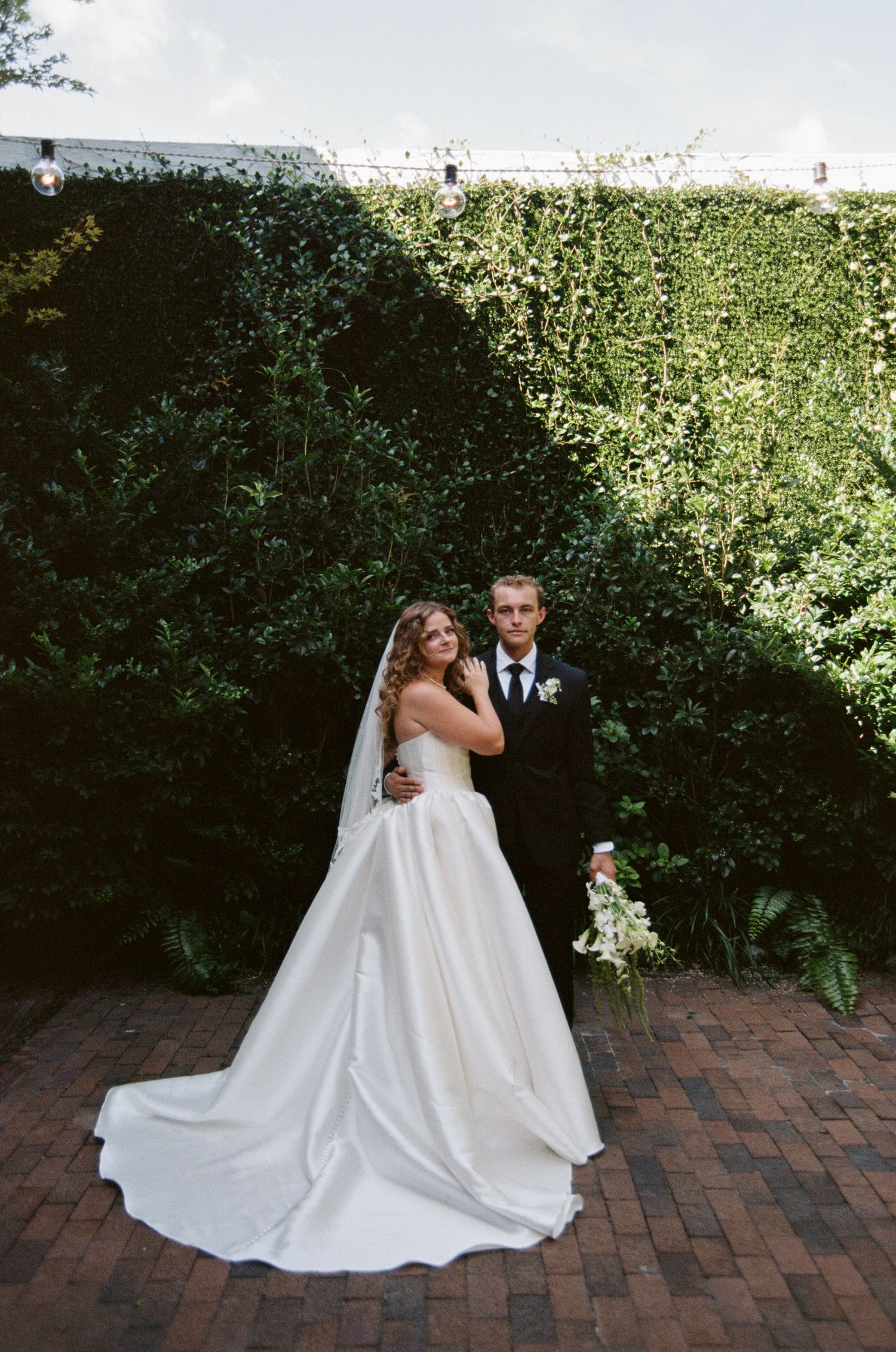 A couple in wedding attire poses together on a brick patio with lush green hedges in the background.