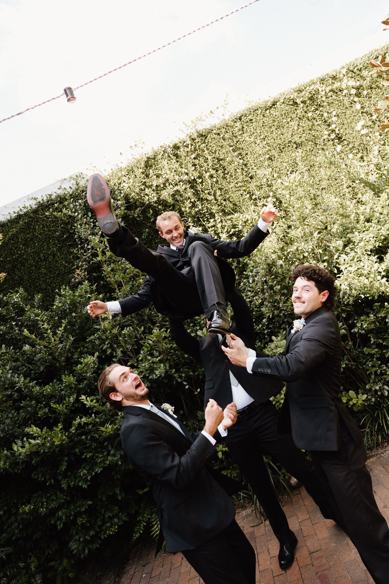 Two people in black suits perform playful martial arts moves on a brick patio with greenery in the background.
