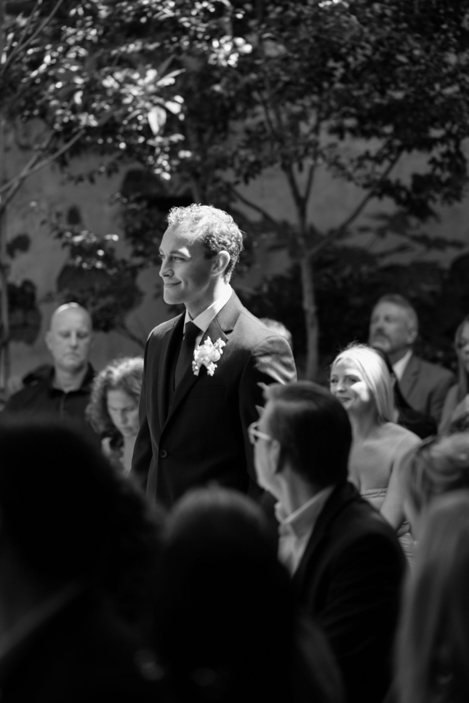 A group of people in formal attire gather outdoors under trees in black and white photography.