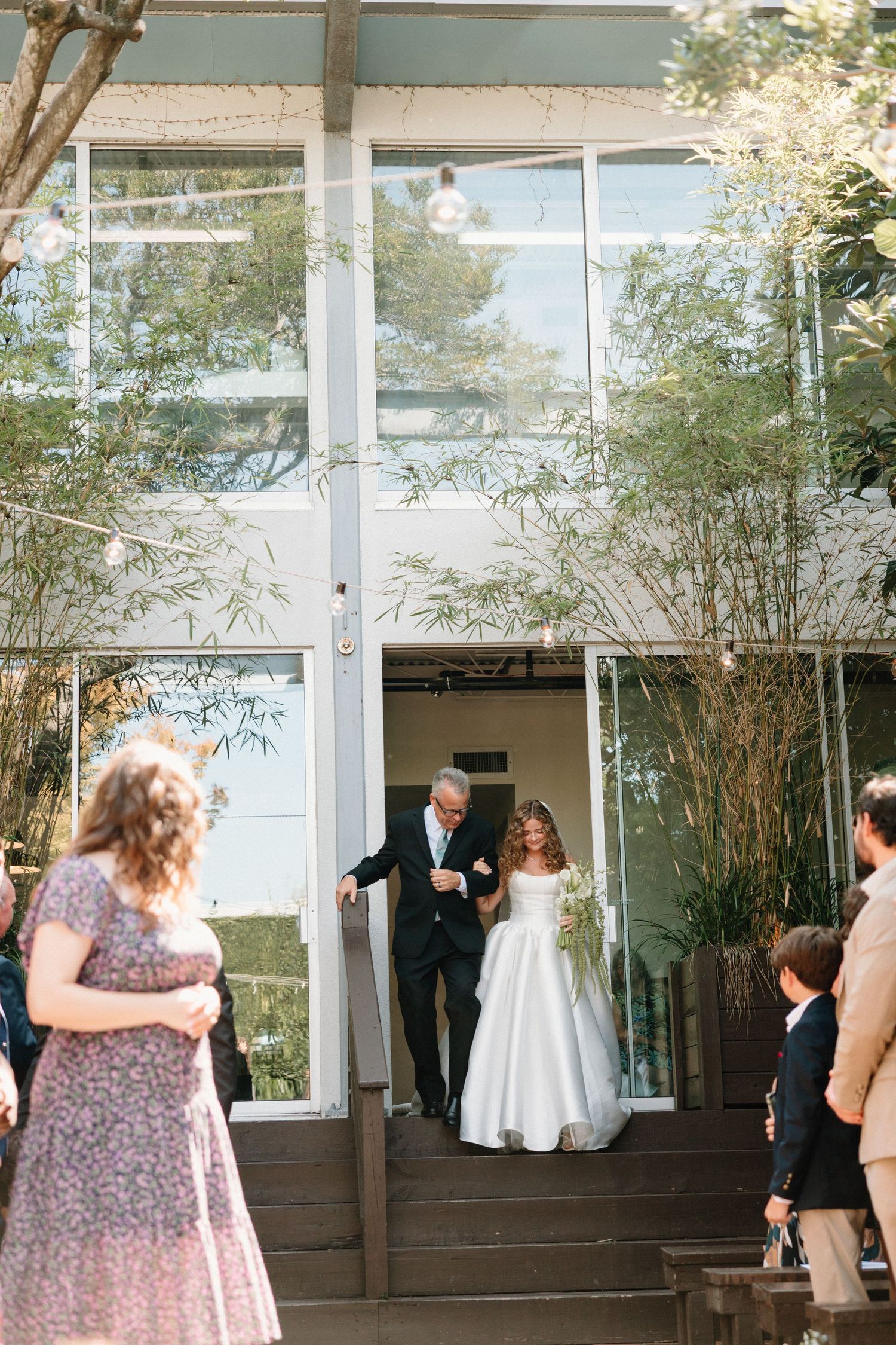 A wedding reception takes place in a modern glass-walled venue surrounded by bamboo plants and natural light.