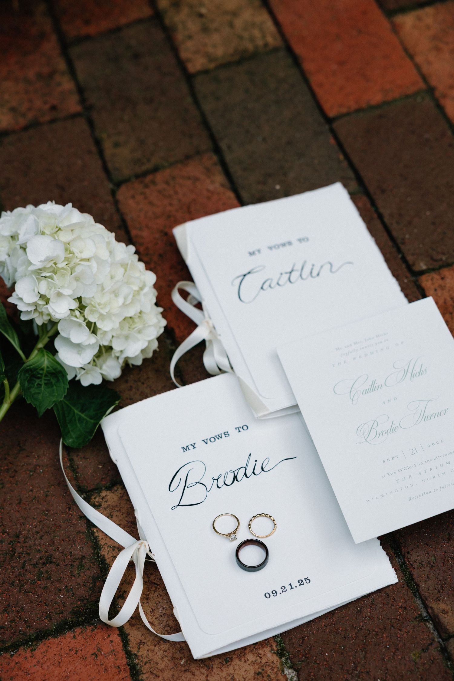 White wedding stationery and gold rings arranged with hydrangea flowers on a brick surface.