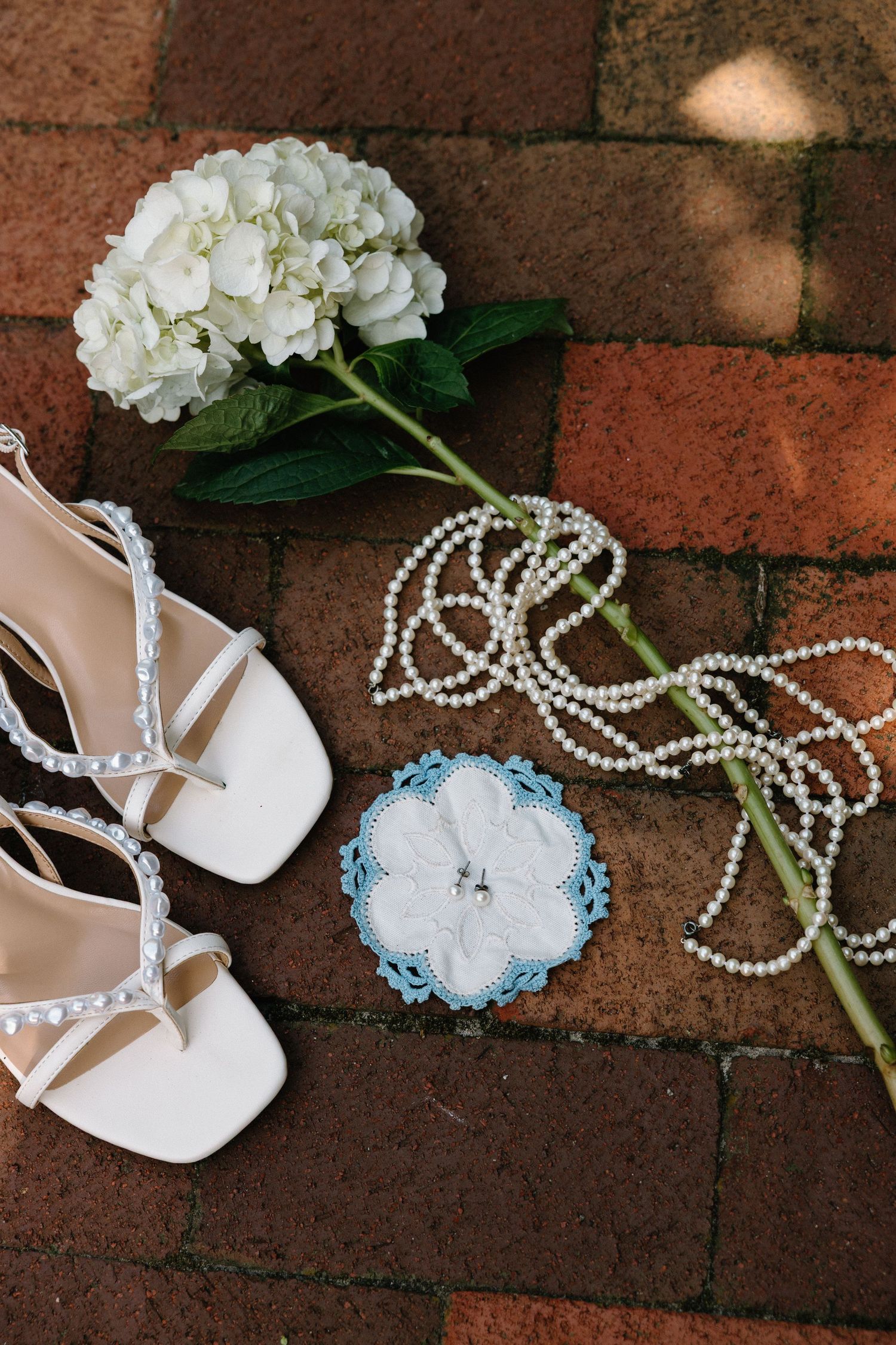 Wedding accessories including white heels, pearl necklaces, blue garter, and white hydrangea flowers arranged on brick pavement.