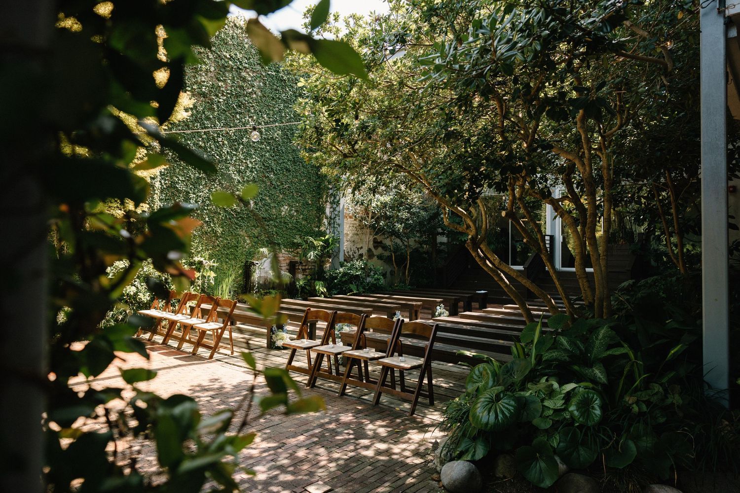 Rustic outdoor wedding venue with wooden chairs arranged in rows under dappled sunlight filtering through trees.