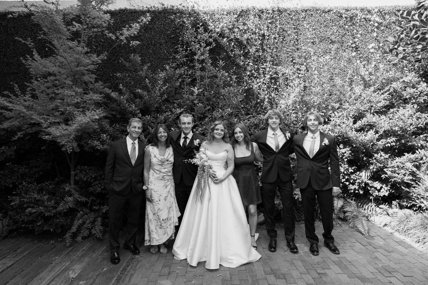 Black and white photo of a wedding party standing together outdoors on a wooden deck with trees in the background.