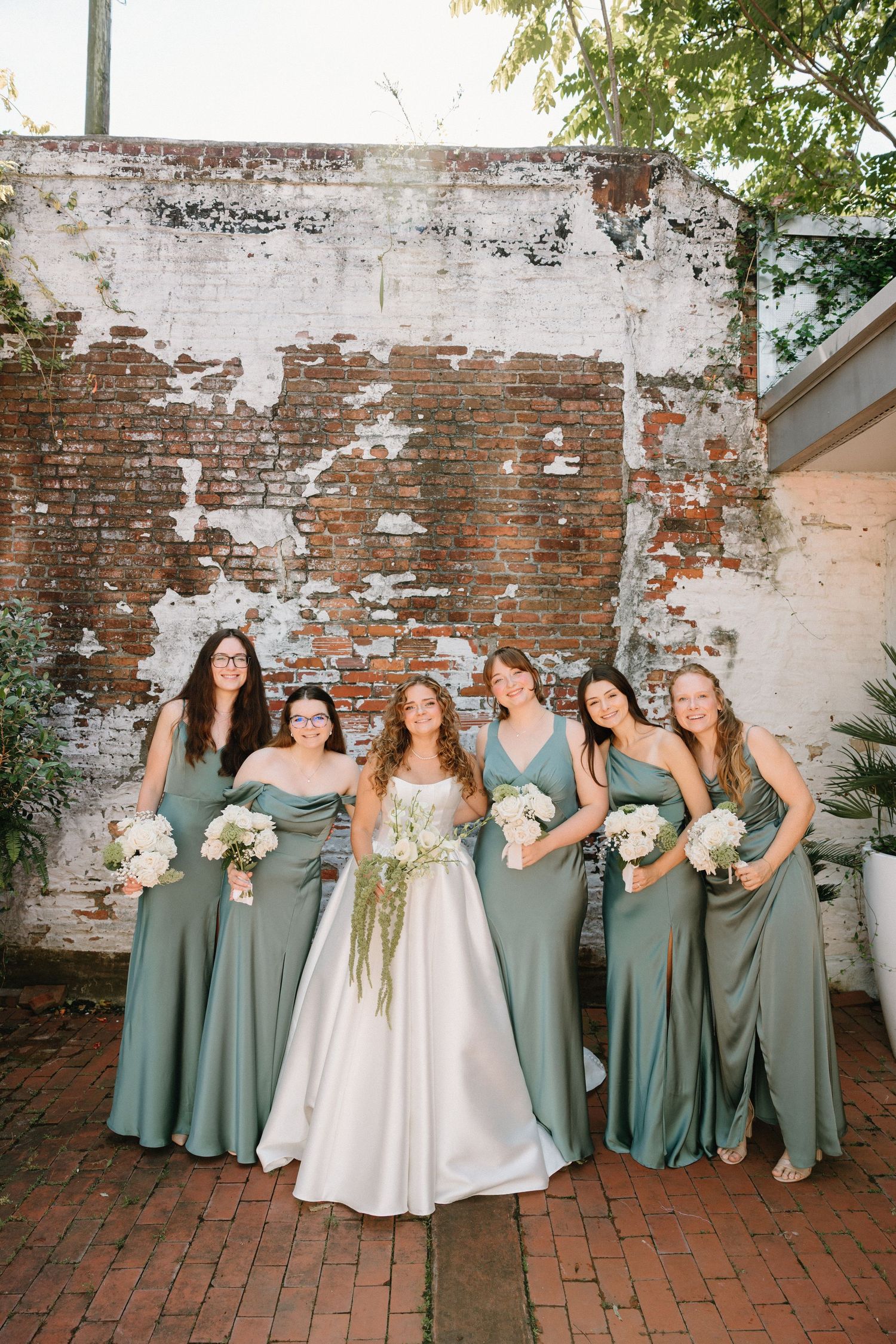 Wedding party in sage green bridesmaid dresses pose together on brick patio against weathered white brick wall.