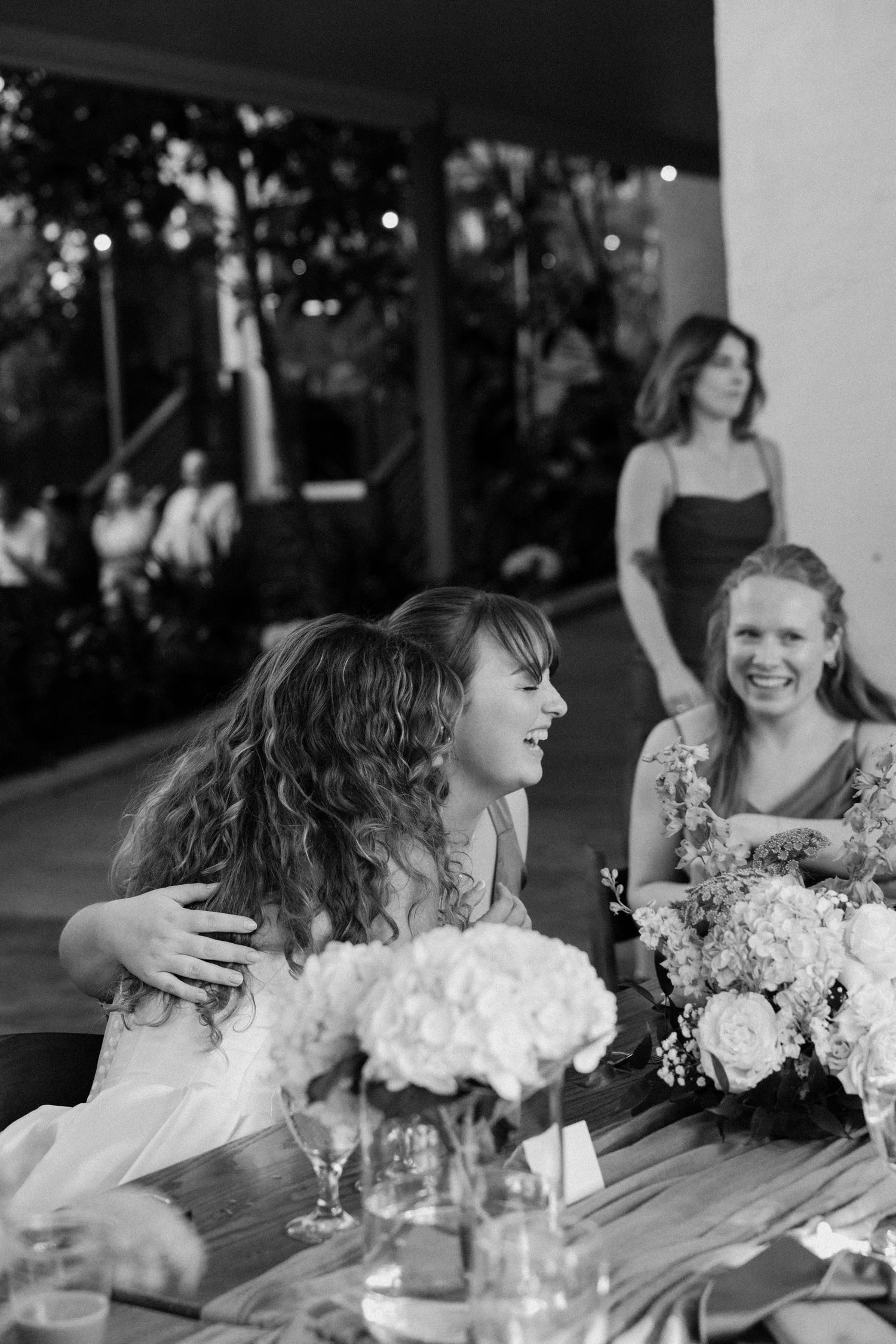 A series of candid black and white photos showing people seated at a wedding reception table with floral centerpieces.