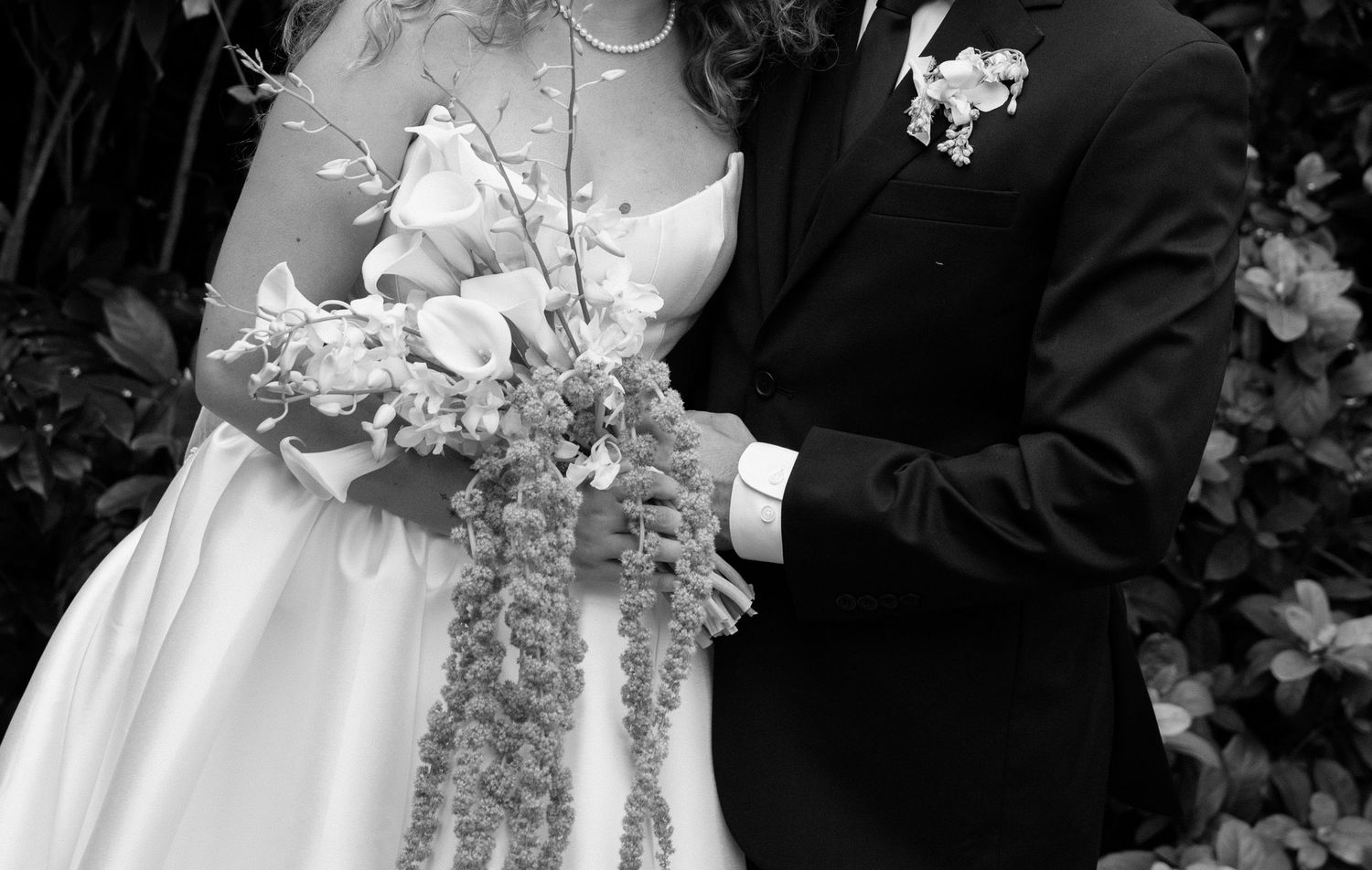 A black and white wedding photograph showing hands holding a cascading floral bouquet with white flowers.