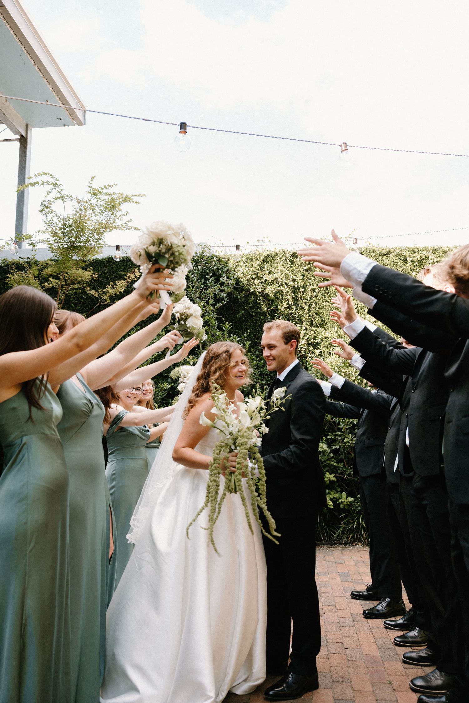 Wedding party celebrates with raised arms on a brick patio as flower petals are tossed into the air.