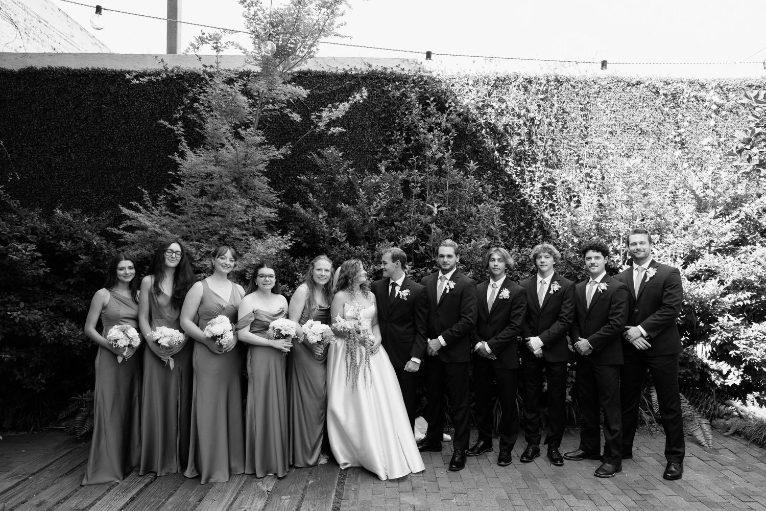 A black and white wedding party photo showing groomsmen and bridesmaids posing together outdoors in formal attire.