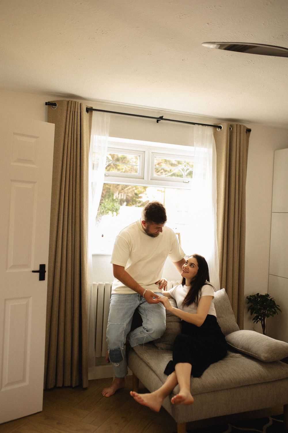 Couple sharing tender moments while relaxing together on a gray couch in a sunlit living room.