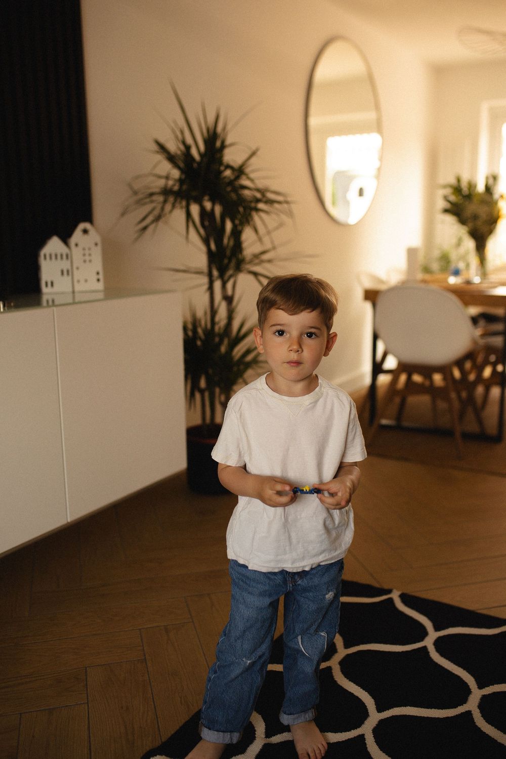 A child in a white t-shirt and jeans plays in a minimalist living room with indoor plants and hardwood floors.