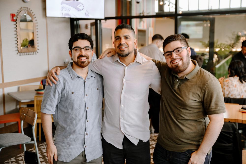 A group of people posing together in a casual restaurant setting with white walls and wooden furniture.