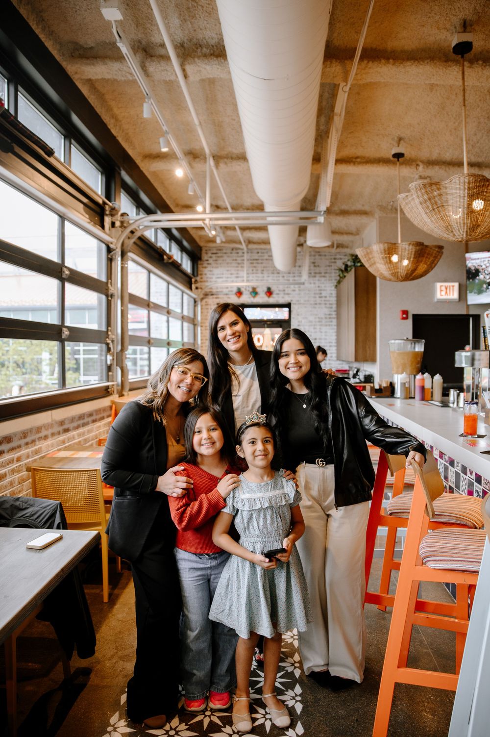 A group poses together at a casual restaurant with exposed industrial ceiling, large windows and orange chairs.
