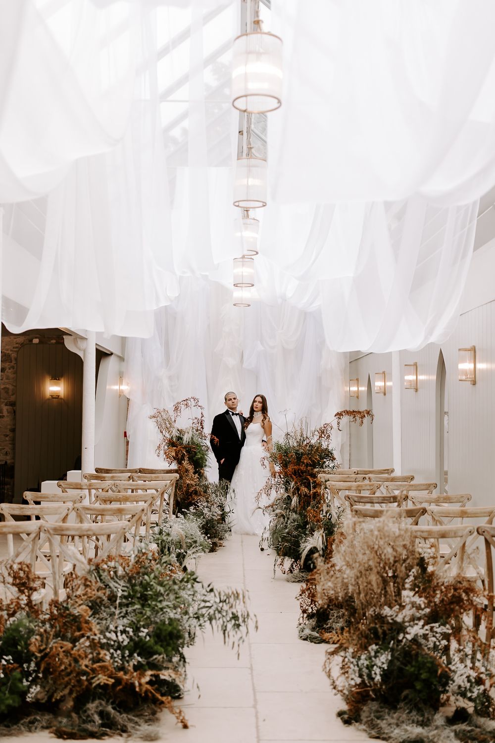 A wedding ceremony space with white draping and romantic floral arrangements lining the aisle.