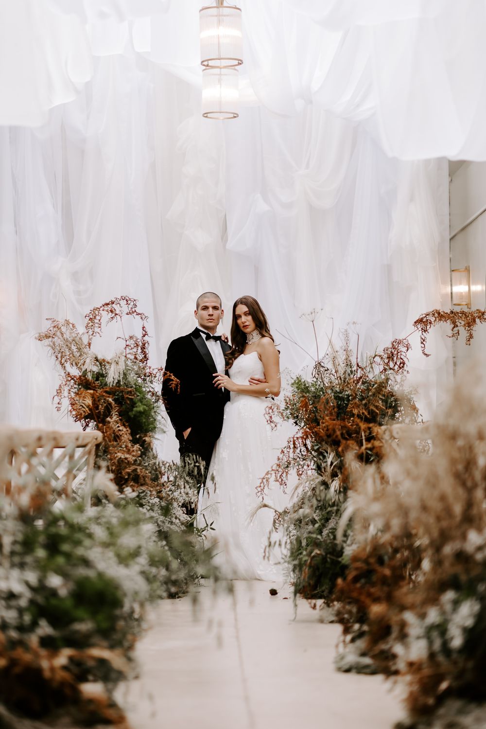 A wedding couple stands in an elegant venue with white draping and autumn-toned floral arrangements lining the aisle.