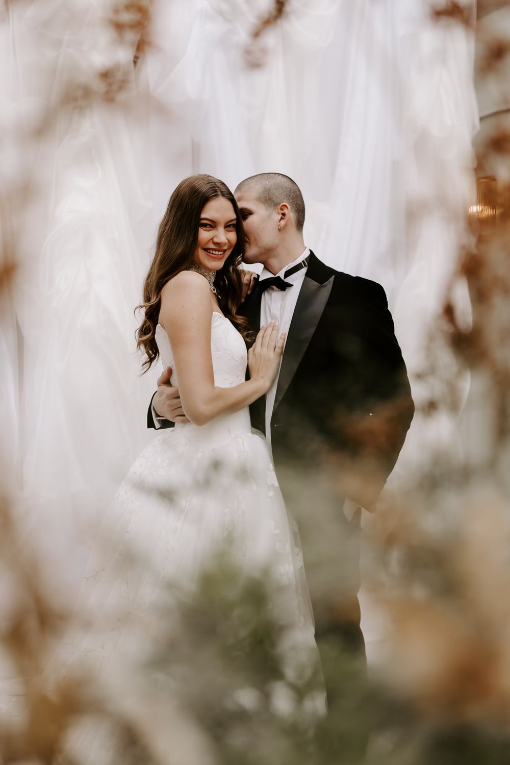 A romantic wedding couple embraces outdoors in soft focus with dreamy bokeh effect through autumn foliage.
