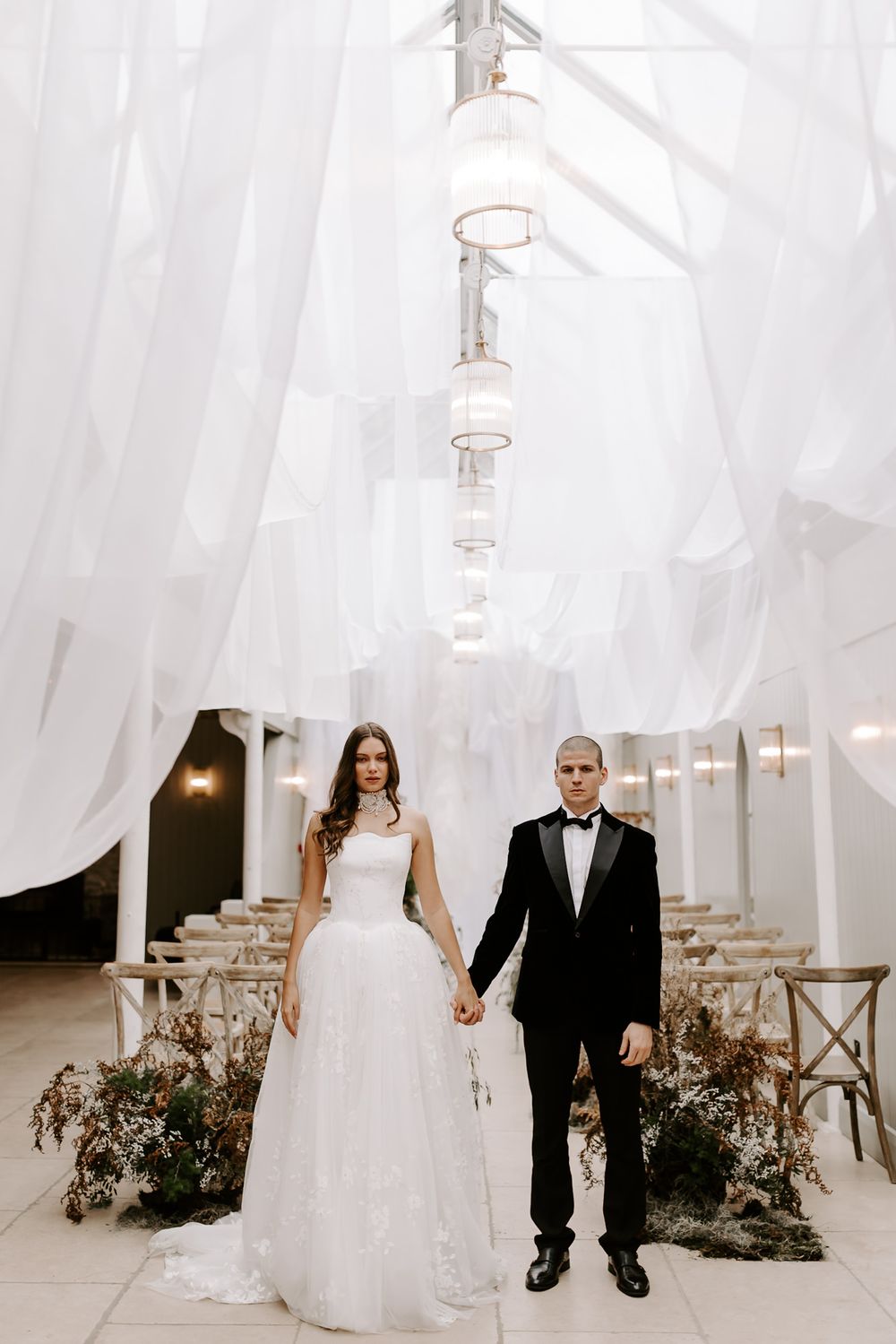 A couple stands together in an elegant white wedding venue decorated with flowing fabric and lanterns.