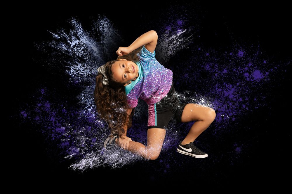 Young girl striking a dynamic hip-hop dance pose with purple and white powder explosion on black background.