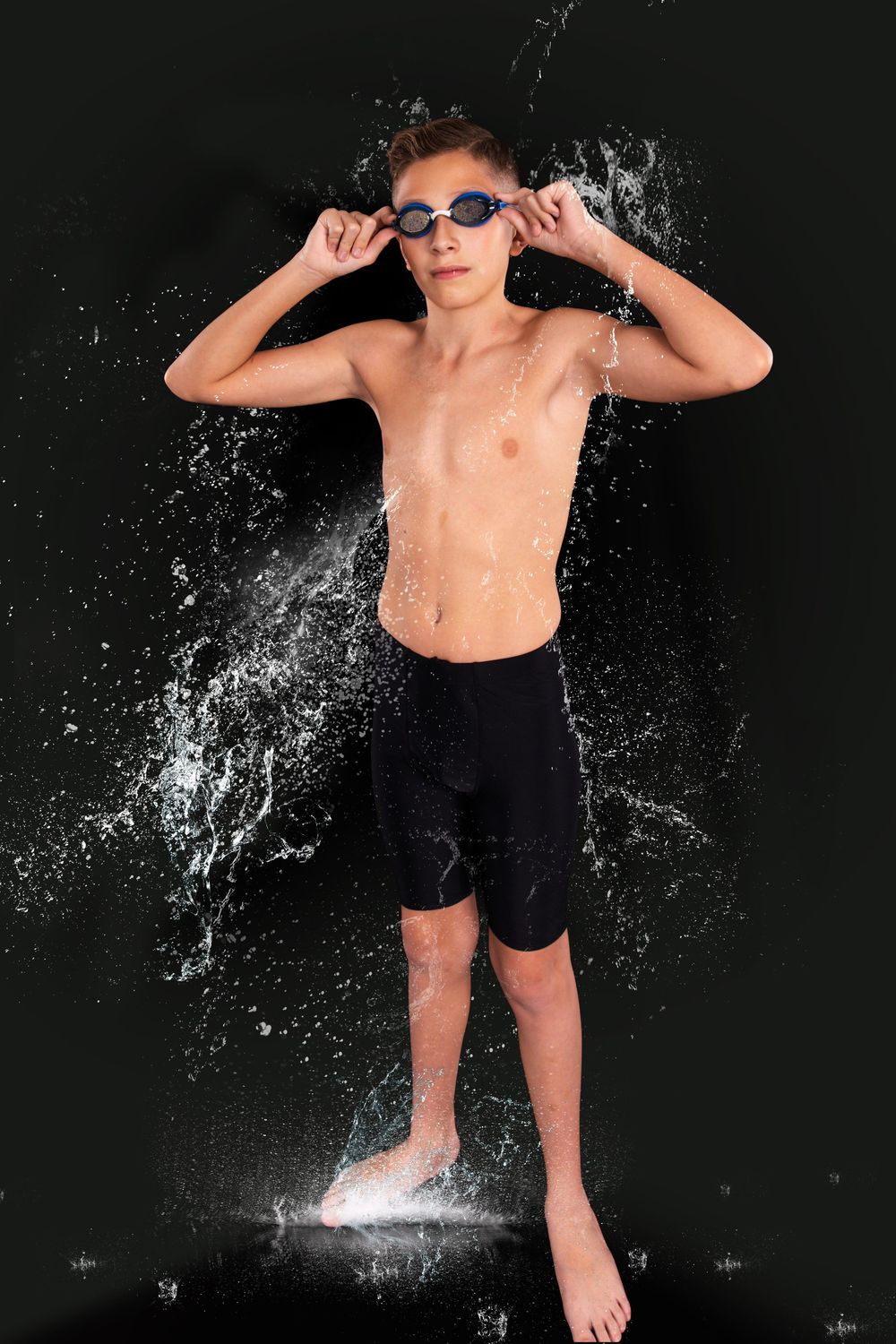 Young male swimmer in black shorts and goggles poses with water splashing around him on dark background.