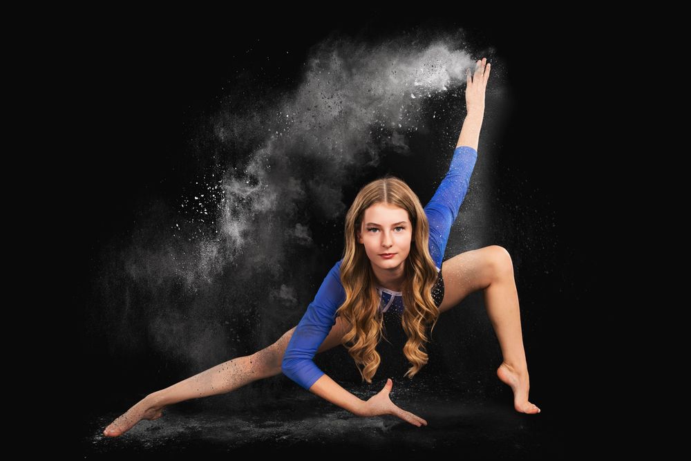 Flexible gymnast in blue leotard performs splits on dark stage with white powder dust cloud effect.