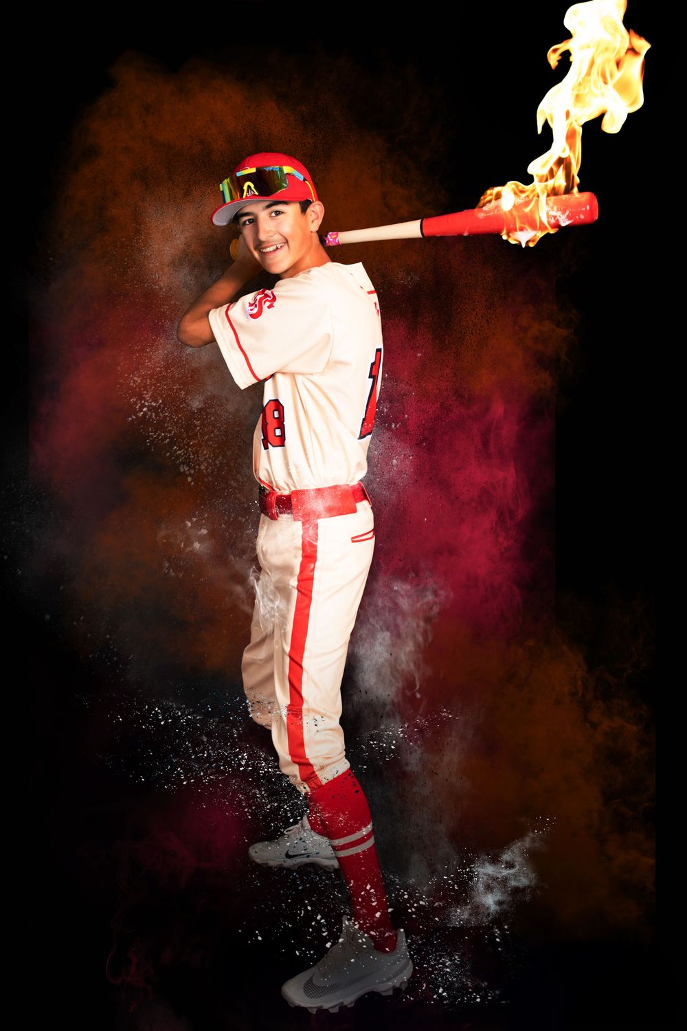 Baseball player in red and white uniform poses with a flaming bat, surrounded by colorful powder explosion on black background.