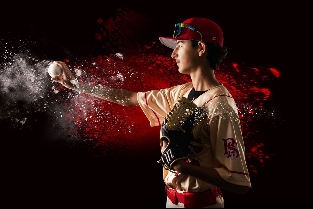 Young baseball pitcher in red uniform mid-throw with dramatic red powder explosion effect on dark background.