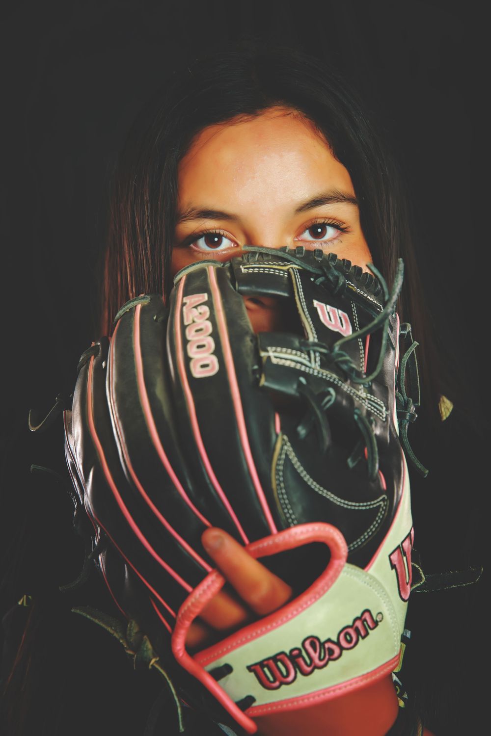 A person holds up a black and pink Wilson baseball glove to cover their face against a dark background.