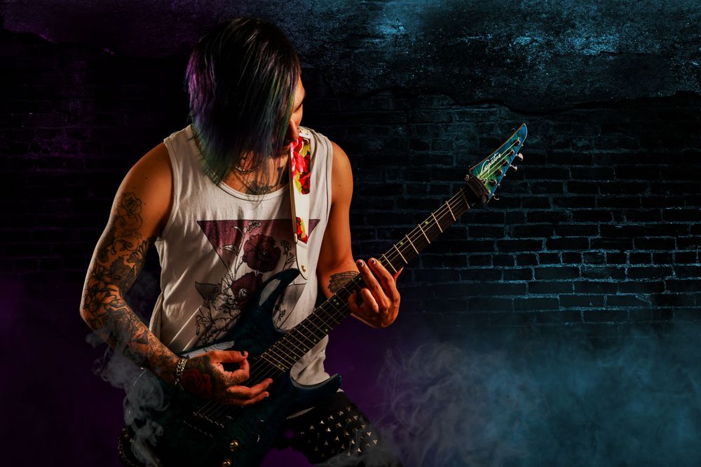 Tattooed musician in a sleeveless shirt plays electric guitar with dramatic purple stage lighting against dark brick wall.