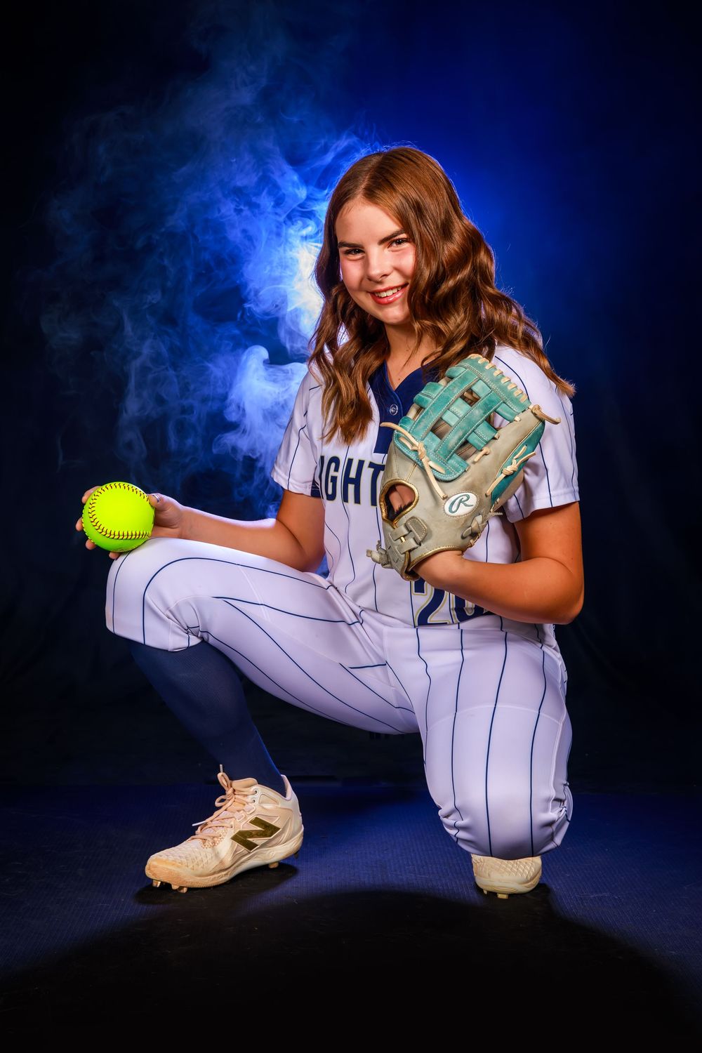 A softball player in a white pinstriped uniform poses with a glove and ball against a dramatic blue smoky background.