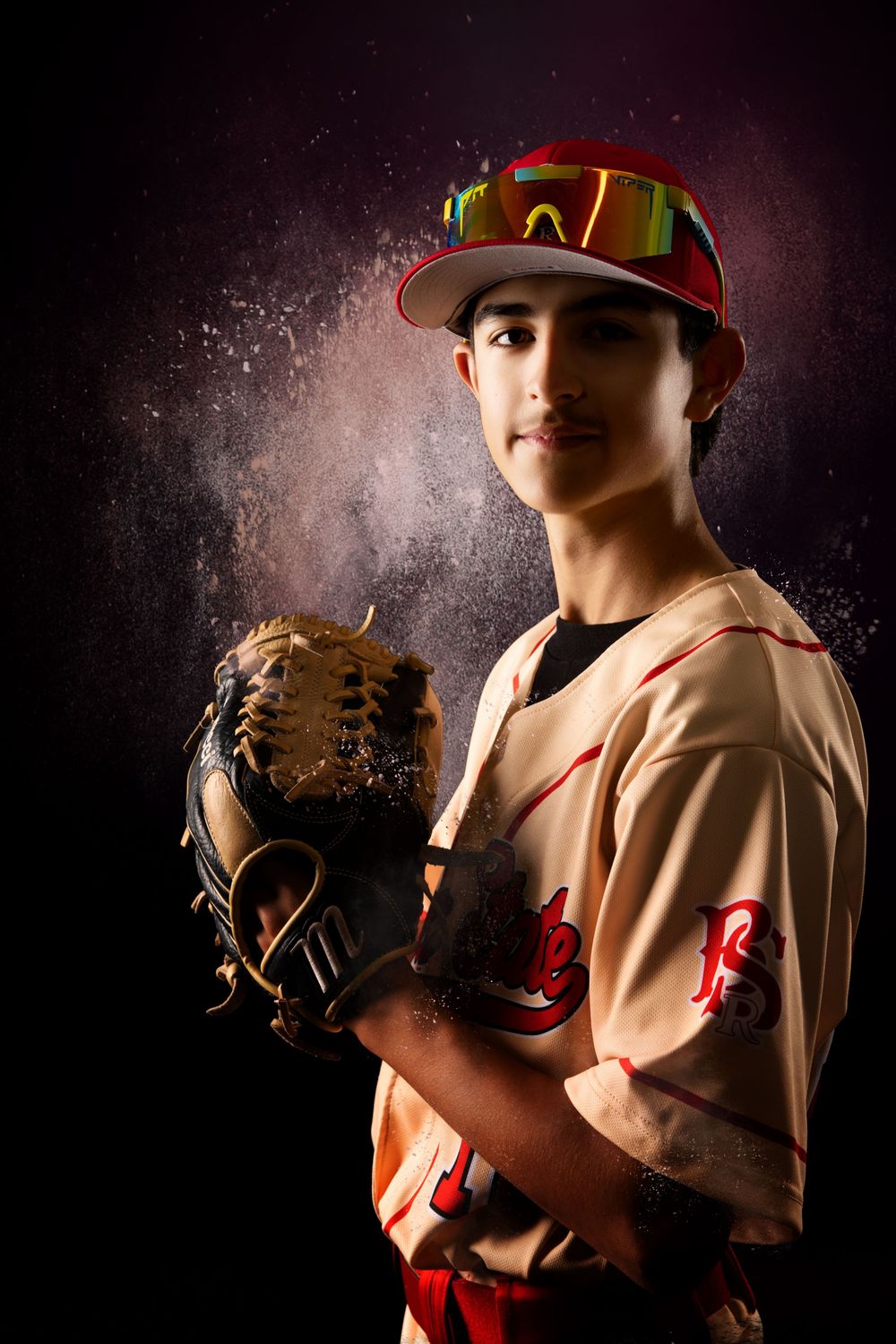 Young baseball pitcher in cream uniform and red cap poses with glove, chalk dust swirling against dark background.