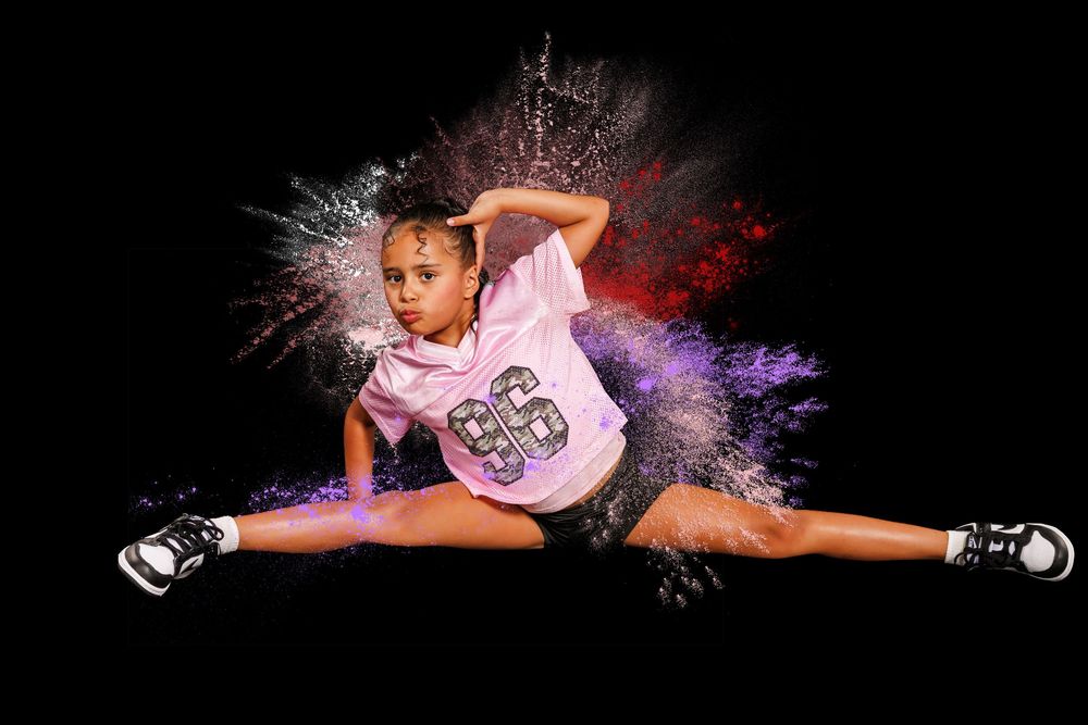 Young dancer mid-split leap in white jersey and shorts with colorful powder explosion on black background.