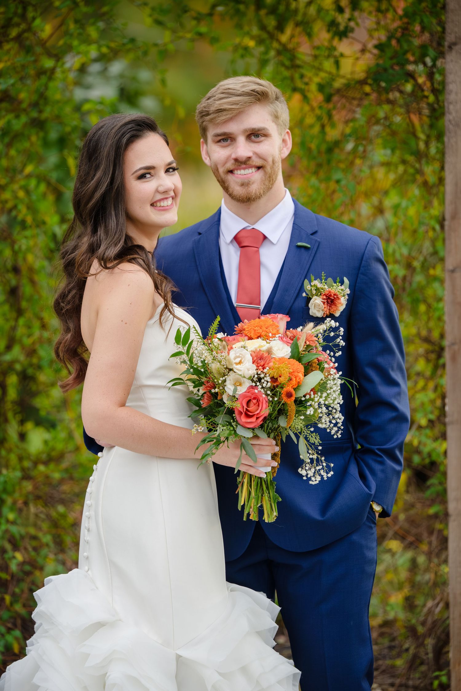 A couple in wedding attire poses outdoors with coral and orange floral bouquets in the autumn at Running Horse Farm in Geneva Ohio