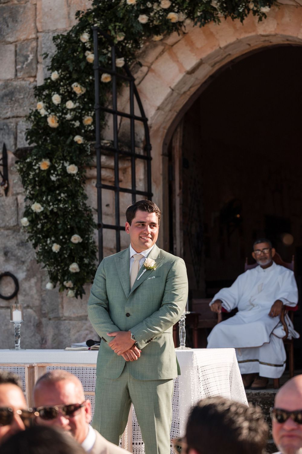A person in a light green suit stands at an outdoor venue with stone archways and climbing roses.