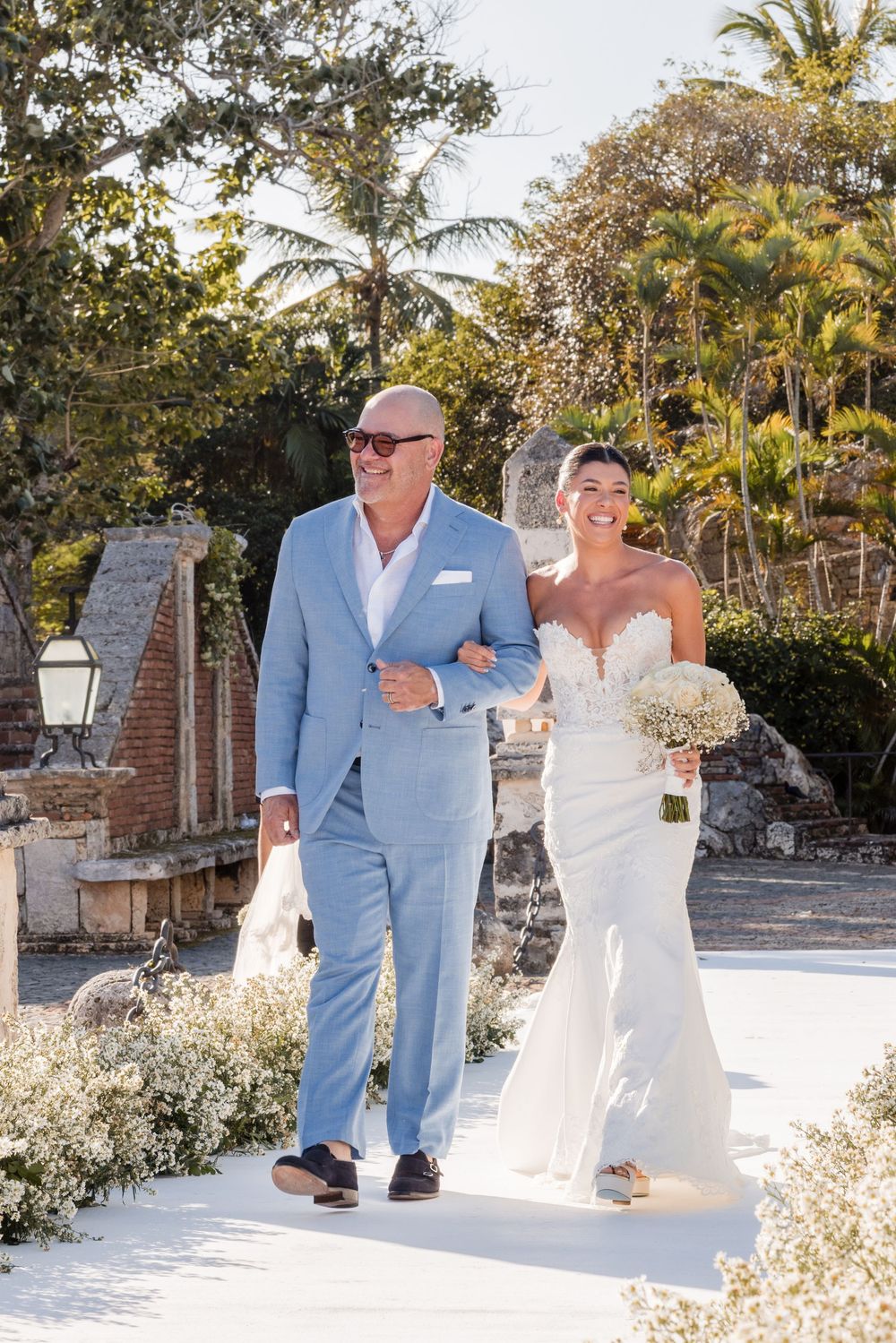Wedding couple in a tropical garden setting with palm trees and white flowers, groom wearing light blue suit.