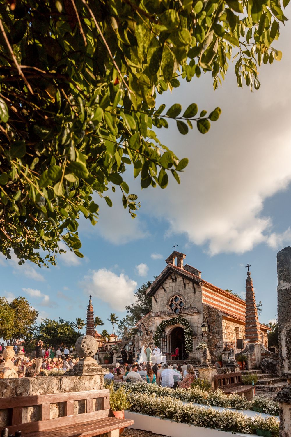 Historic stone chapel with ornate architecture and green foliage overhead on a sunny day with blue skies.