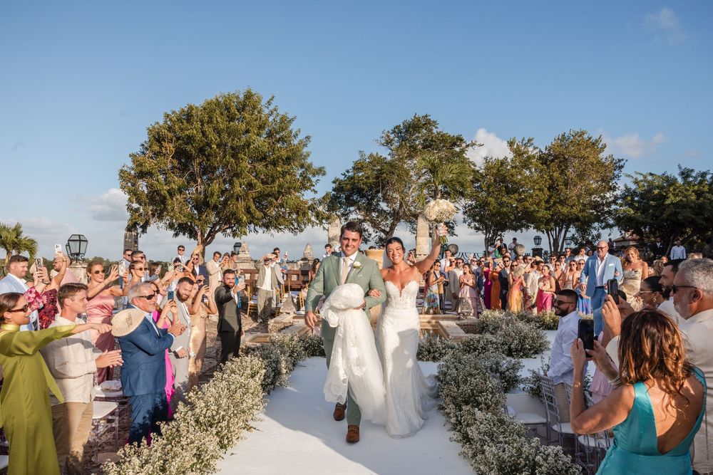 Wedding guests line a white aisle at an outdoor ceremony with palm trees in the background.