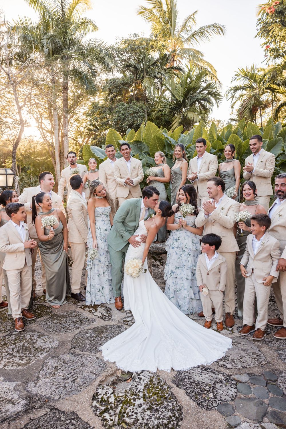Wedding party poses together outdoors among palm trees and stone pathways at sunset.