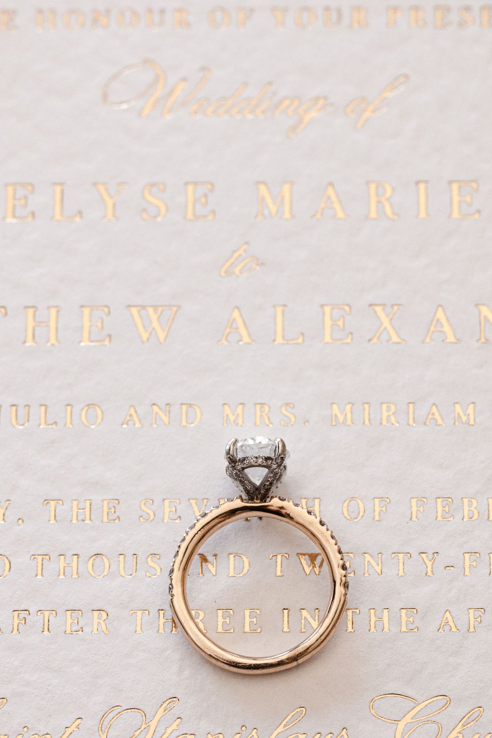 A diamond engagement ring rests on an elegant wedding invitation with gold lettering.