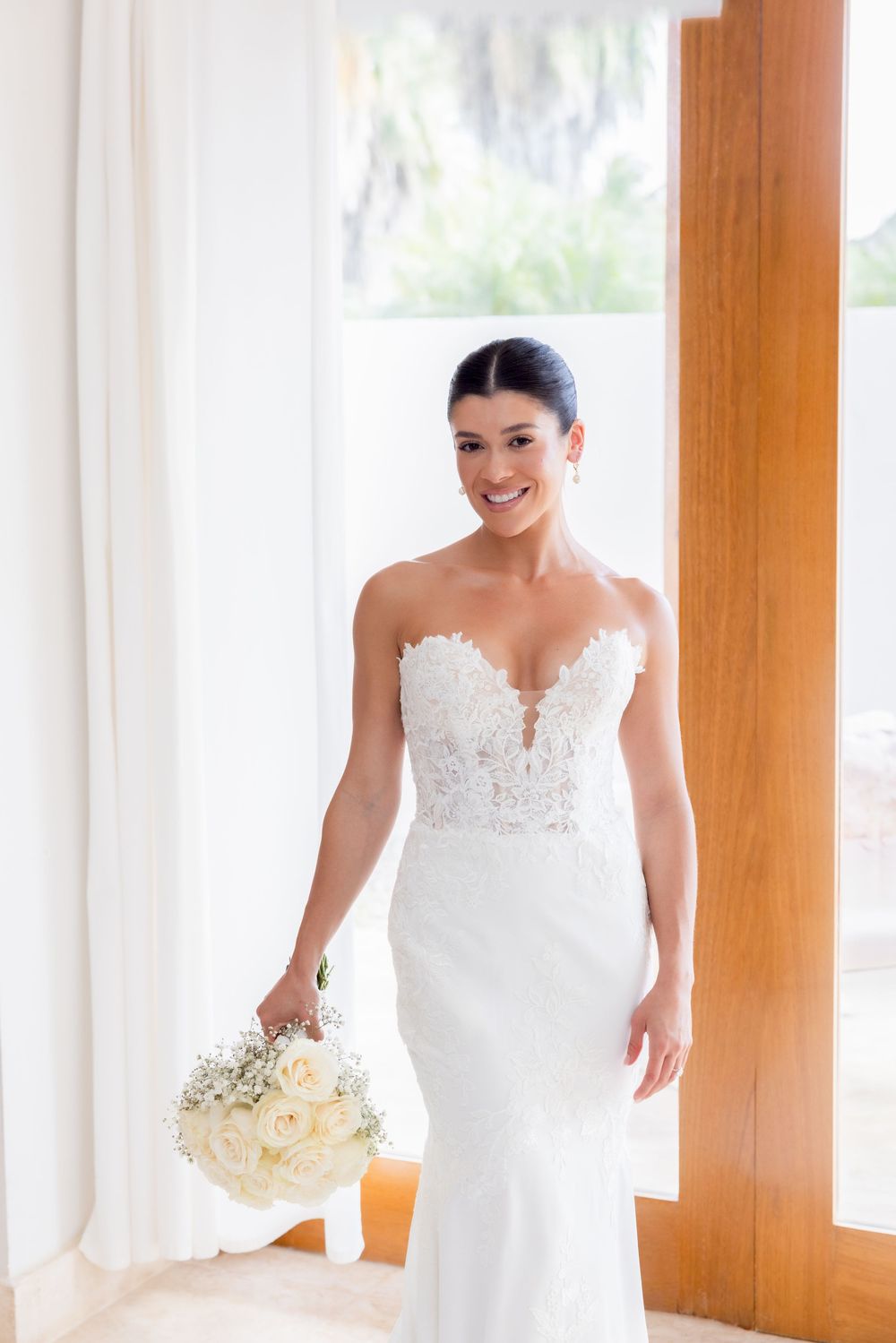 Bride in elegant white wedding gown holds bouquet while standing in sunlit room with wooden pillars.