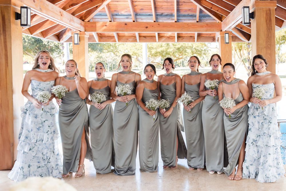 A wedding party in gray bridesmaid dresses and floral print dresses poses under a covered outdoor pavilion.