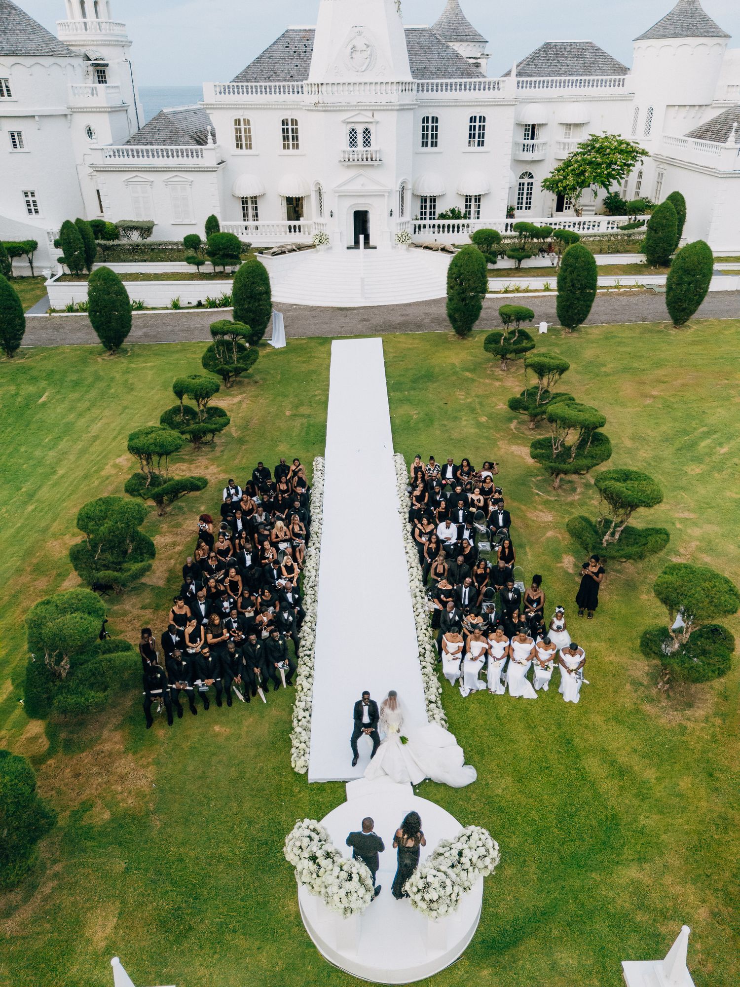 Aerial view of an elegant white wedding ceremony setup with guests arranged in formation on a manicured lawn.