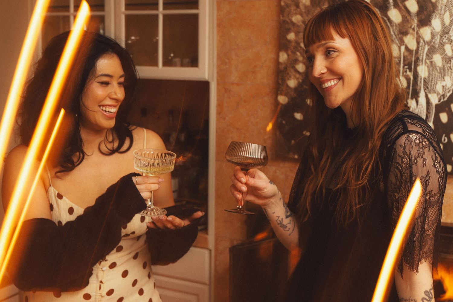 Two friends share drinks and laughter at a warmly lit gathering with orange lighting and festive ambiance.