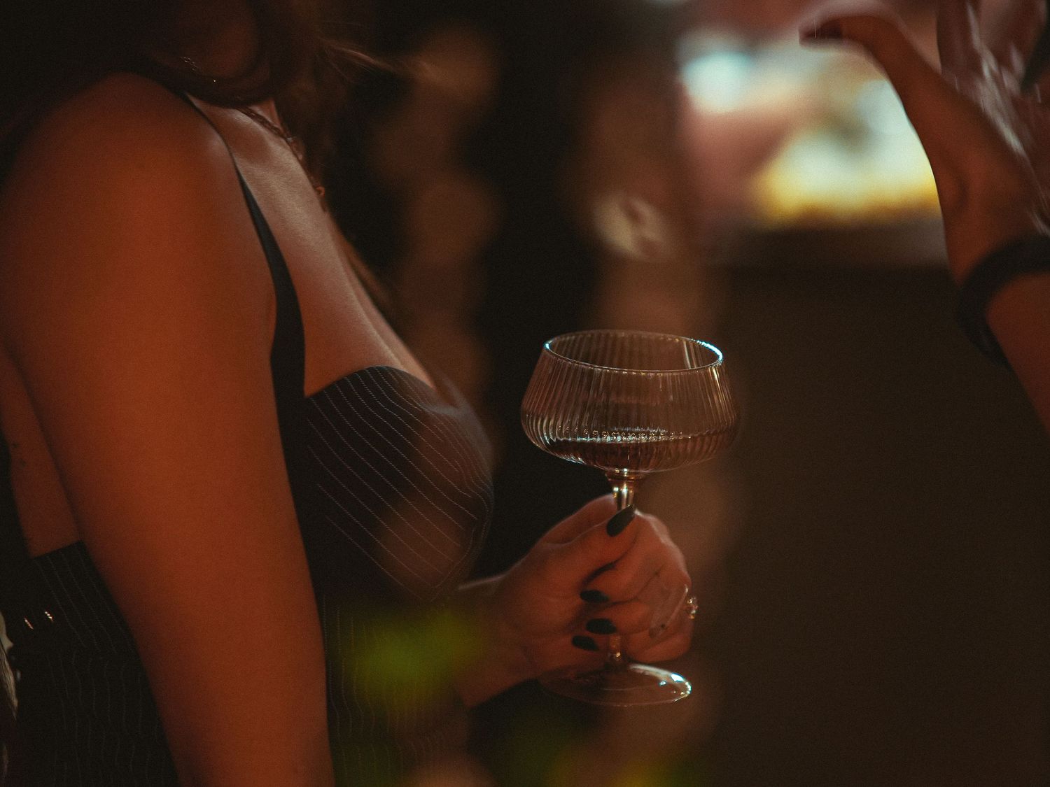 A person holds a coupe glass with a dark beverage in a dimly lit setting with atmospheric lighting.