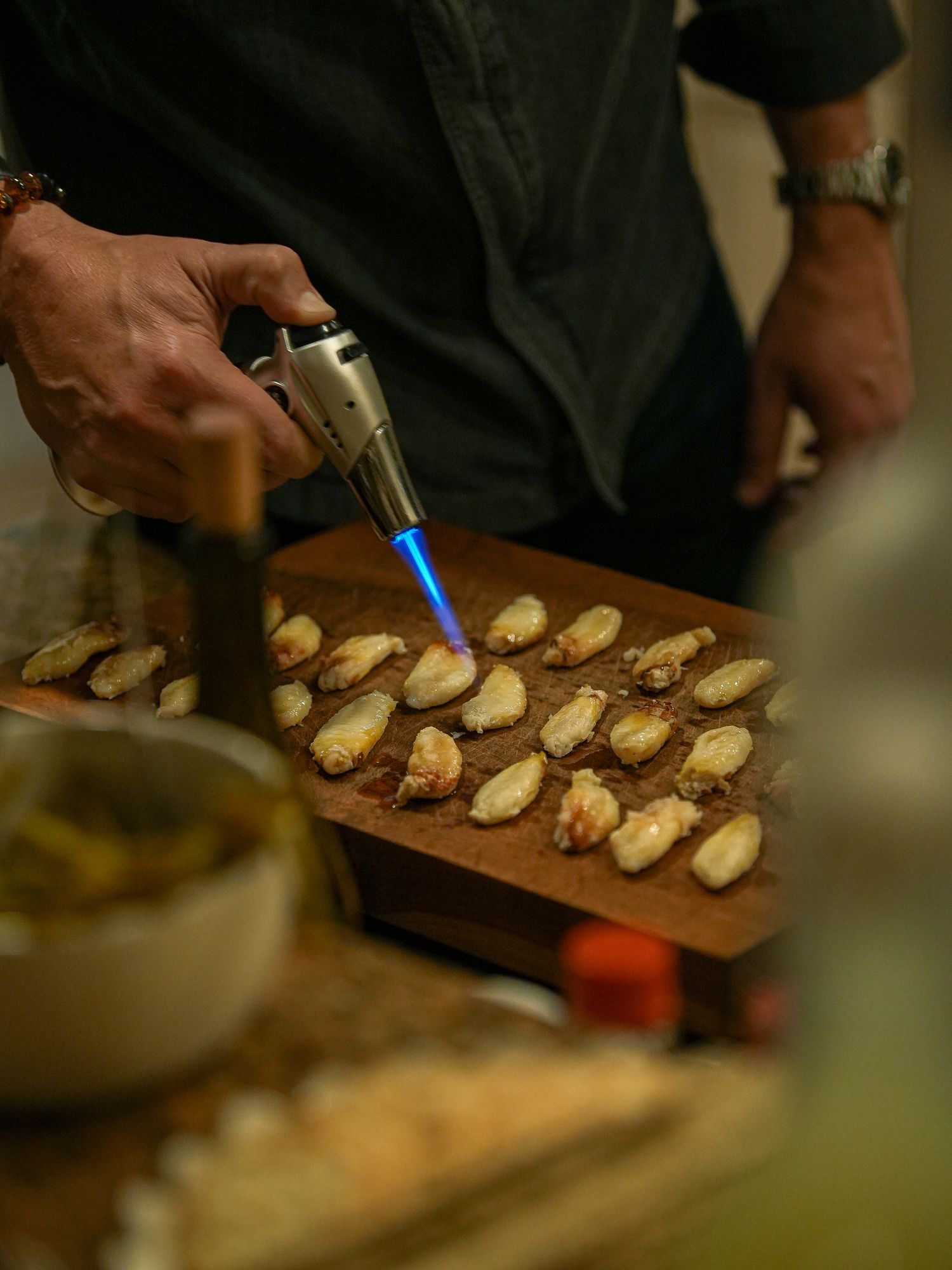 A wooden cutting board displays rows of sliced baguette pieces being prepared with toppings in a kitchen setting.