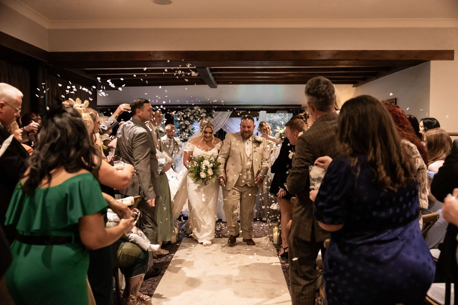 Wedding party guests toss rose petals at newlyweds exiting brick church building in celebratory moment.