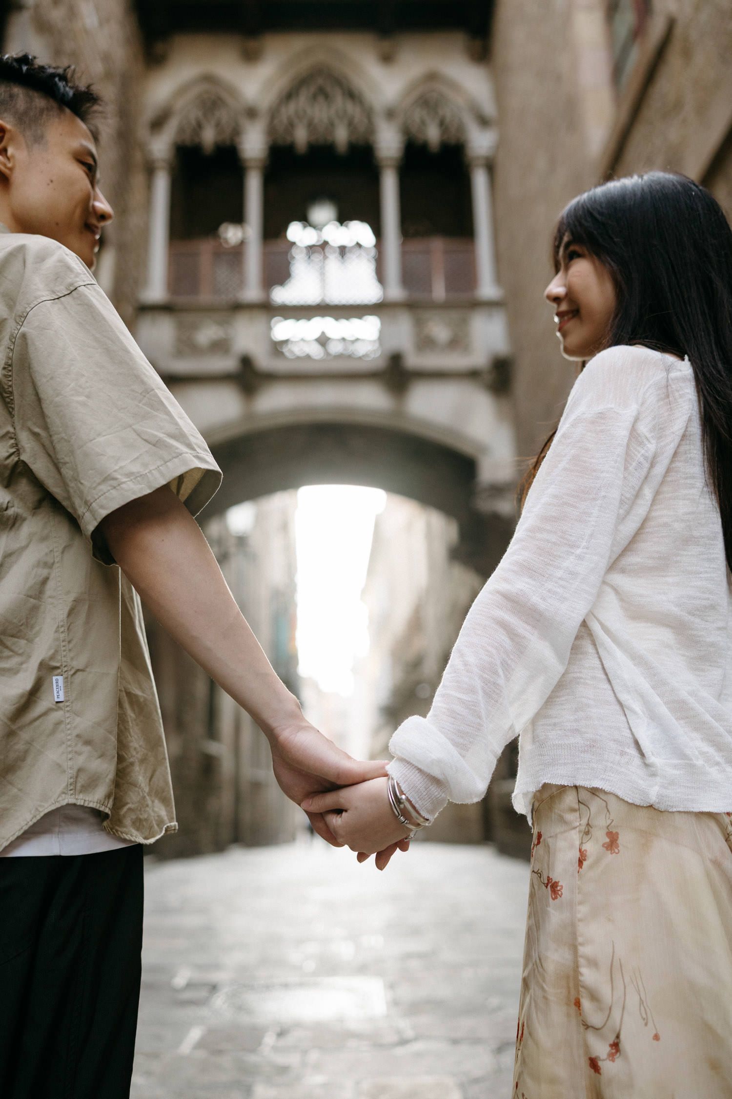 Couple in light colored outfits holding hands in Gothic architectural setting.
