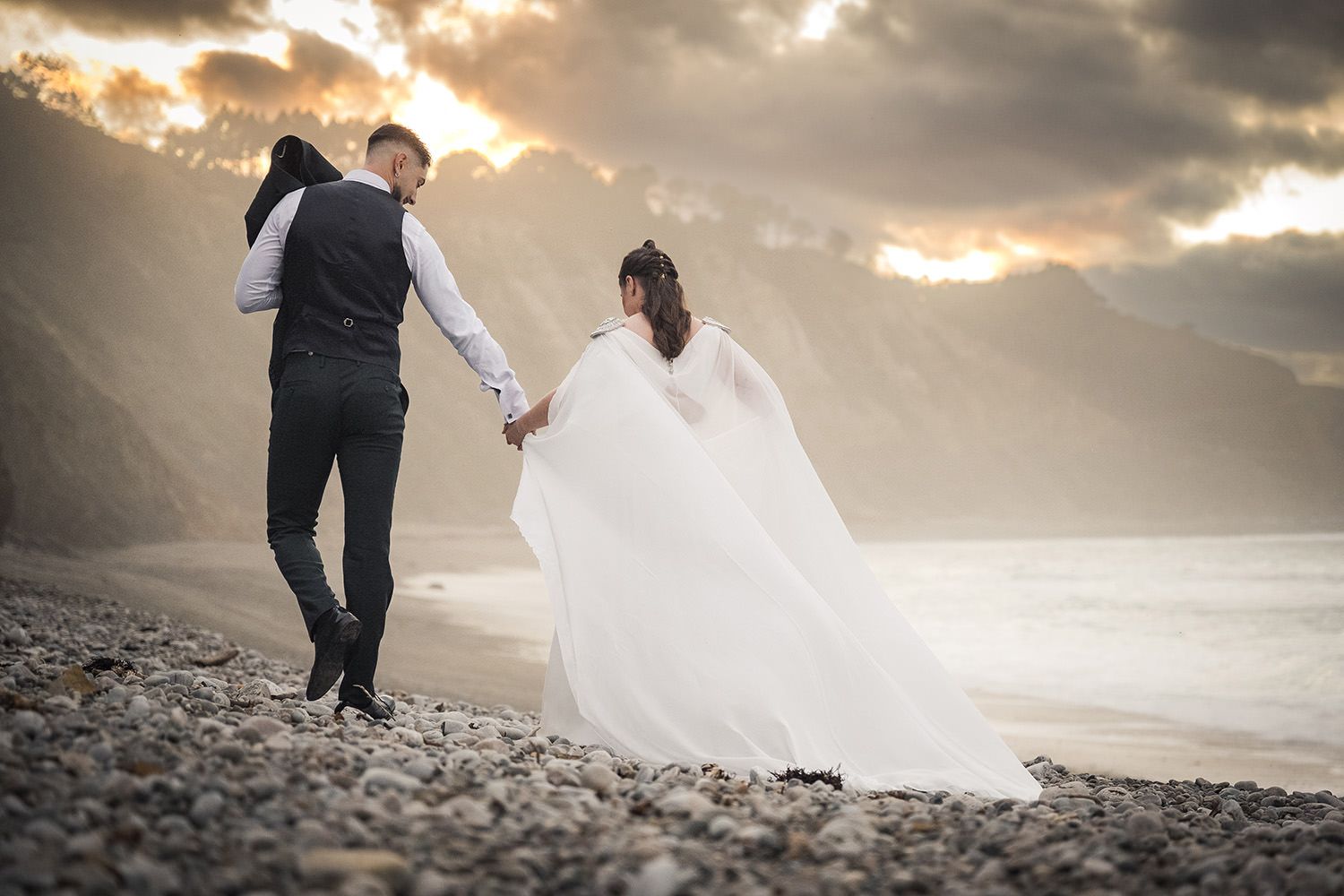 Postboda en Asturias – sesión creativa al aire libre. Novios pasean cogidos de la mano en una playa de Gijón al atardecer