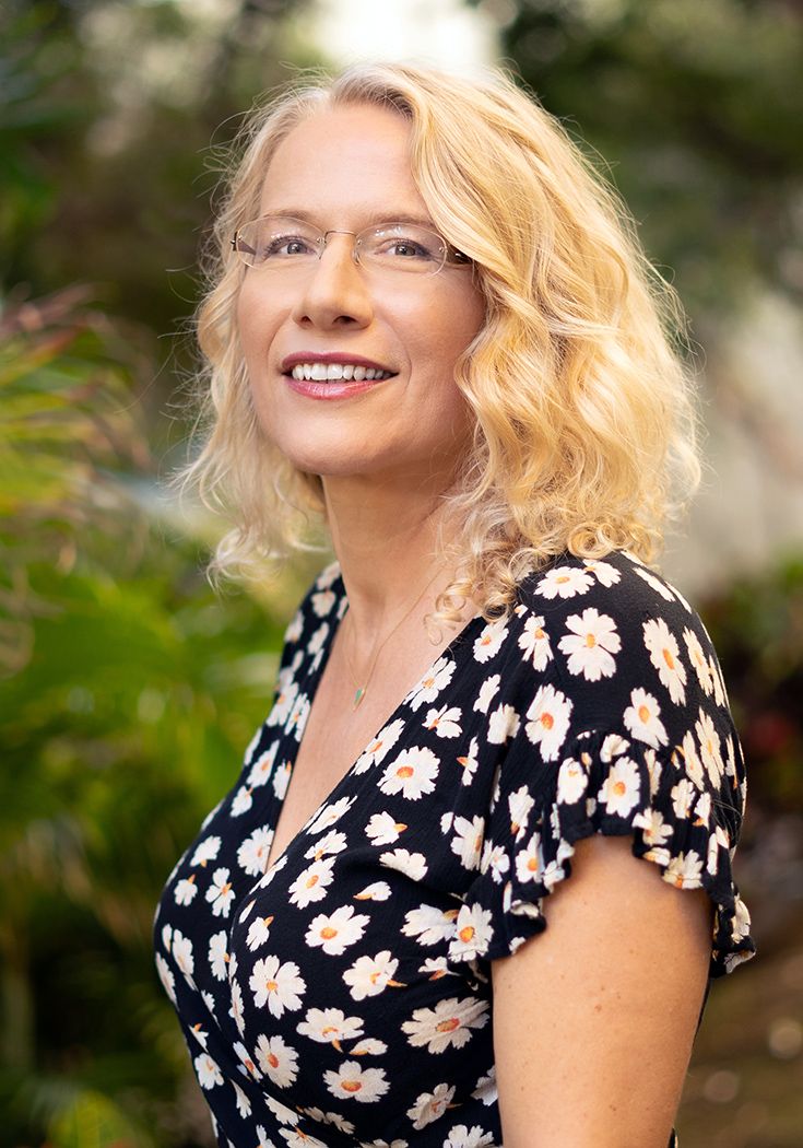 A person wearing a black and white floral dress smiles warmly outdoors with natural greenery in the background.