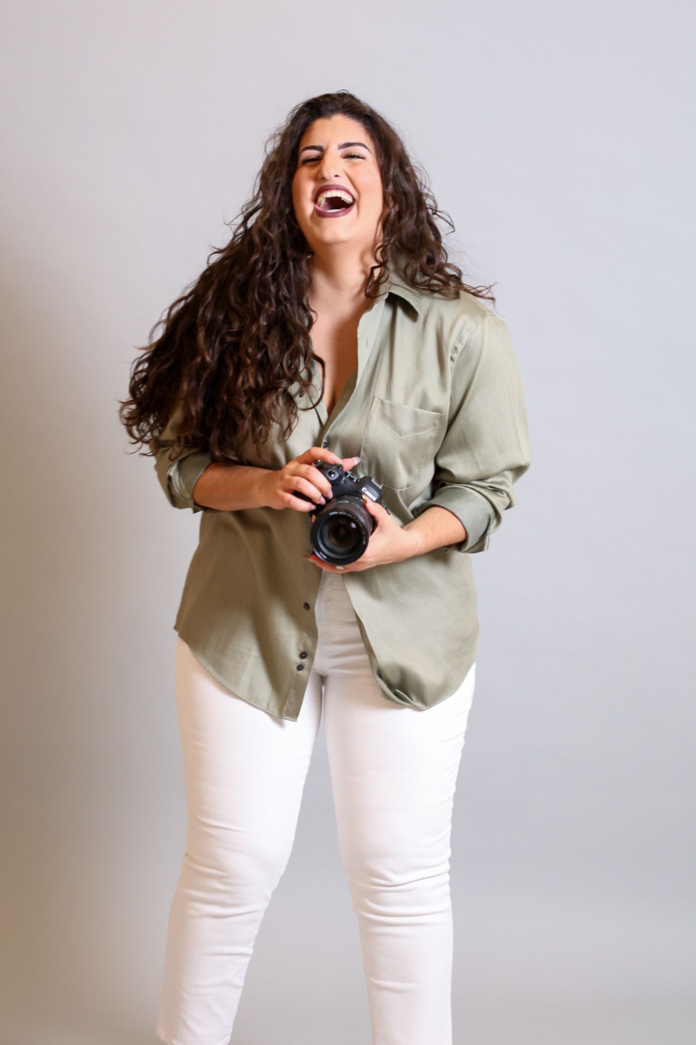 Photographer wearing sage green shirt and white pants laughs joyfully while holding camera against light background.